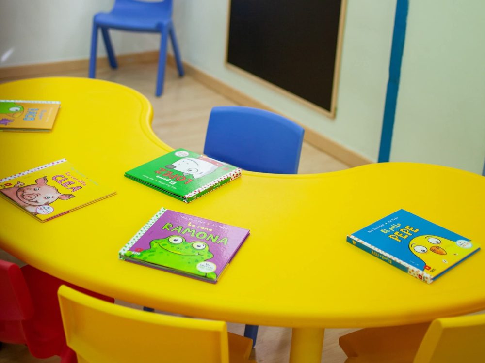 Rincón de lectura con libros infantiles, mesa amarilla y sillas azules en CEI Caracola Utrera.